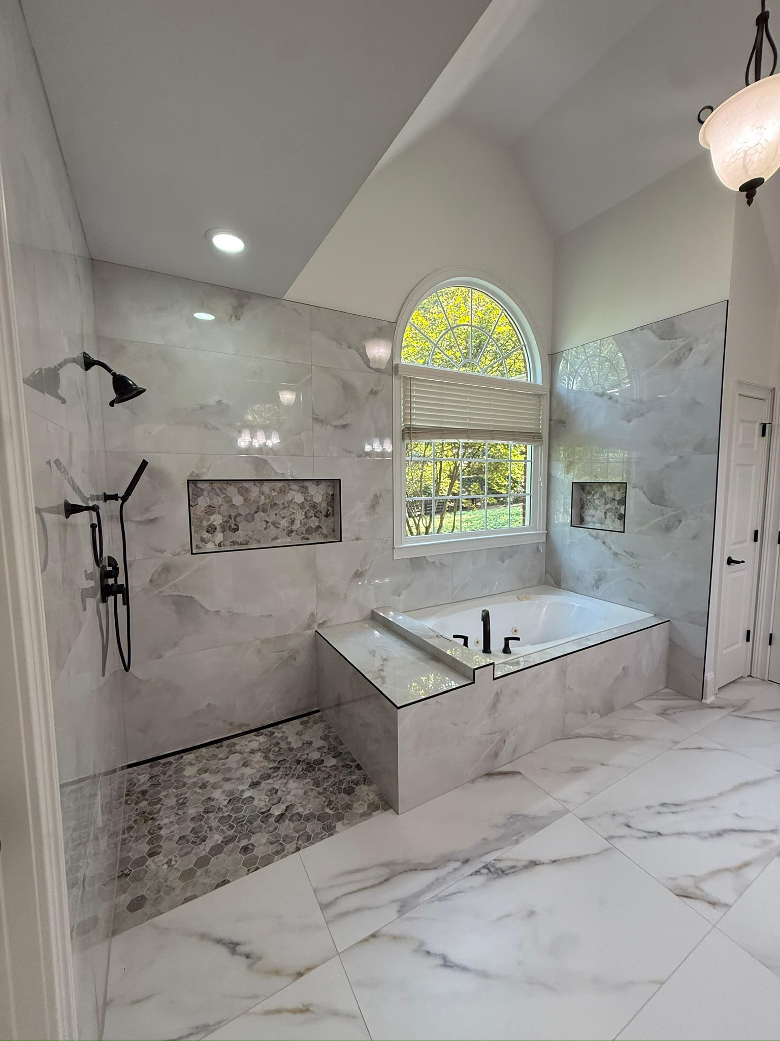 Bathroom with arched window, tub and marble-look wall and floor tile.